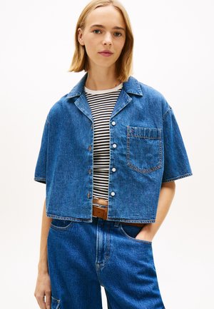 Ung kvinde med kort blondt hår iført en cropped denimjakke med korte ærmer over en sort-hvid stribet skjorte og højttaljede blå jeans.