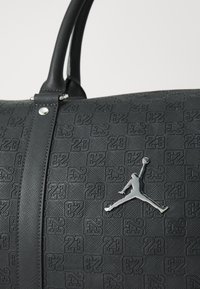 Bolsa de viaje negra con una superficie texturizada que presenta patrones en relieve de "23". Incluye un logo Jumpman plateado y asas negras resistentes.