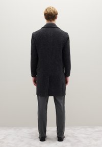 Cappotto in lana grigio scuro con trama lavorata, maniche lunghe, ampio colletto e spacco posteriore, abbinato a pantaloni grigio chiaro e scarpe nere.