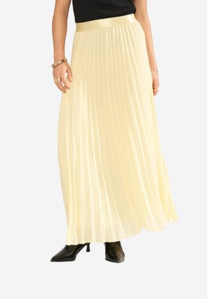 Kvinde iført en lang, lysegul plisseret nederdel, sorte ankelstøvler med spidse tæer og en sort top, der viser smykker på hænderne.