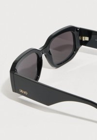 Melni saulesbrilles ar biezu, leņķisku rāmi un tumšām lēcām. Uz sānu daļām zelta krāsā redzams "LIU-JO" logotips. Spīdīgs pārklājums un stabils dizains.