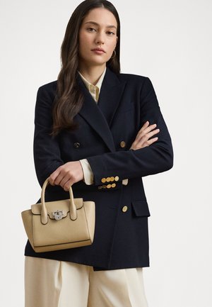 Femme aux longs cheveux foncés, portant un blazer bleu marine à boutons dorés et un pantalon crème, tenant un petit sac à main beige sur un fond uni.