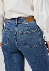 Korkeavyötäröiset siniset farkut, joissa on tekstuuripinta. Farkuissa on takataskut ja vyötäröllä ruskea nahkalappu.