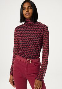 Langarm-Turtleneck-Oberteil mit schwarzem Hintergrund, der ein rotes Herzmuster zeigt. Kombiniert mit roten Cordhosen und einem goldenen Gürtel.