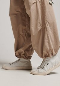 Beige, löst sittande byxor med samlade fållar, tillsammans med vita högkartiga sneakers med texturerad ovansida och grå snören.