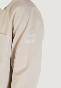 Beige long-sleeve shirt with a chest pocket and a small white patch with "BOSS" embossed on the upper sleeve.
