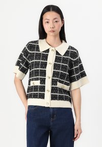 Black and white checkered knitted cardigan with short sleeves, large cream collar, and decorative buttons, featuring two front pockets.