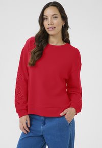 Mujer con cabello largo castaño lleva un suéter rojo con mangas bordadas y vaqueros azules, de pie con una mano en el bolsillo.