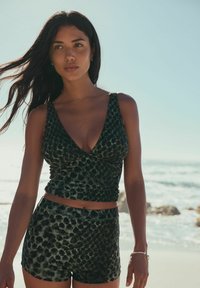 Kvinde med langt mørkt hår iført en mørk ærmeløs top med leopardprint og shorts, der står på en solrig strand med bølger i baggrunden.