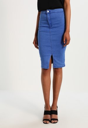 Kvinde iført en blå denim pencilskørt med frontslids, sort top og sorte åbne højhælede sandaler, stående mod en ensfarvet baggrund.