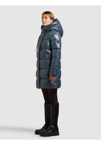 Mörkblå vadderad parkas med blank finish, utrustad med stor huva, dragsko och sidfickor. Bärs med knähöga svarta stövlar.