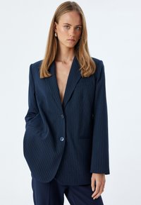Blazer a righe navy in un design strutturato con un solo bottone, caratterizzato da revers a freccia e tasche laterali. Tessuto dalla texture liscia.