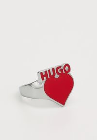 Silver ring featuring a raised red heart with "HUGO" in bold letters. The design is playful with a smooth texture and shiny finish.