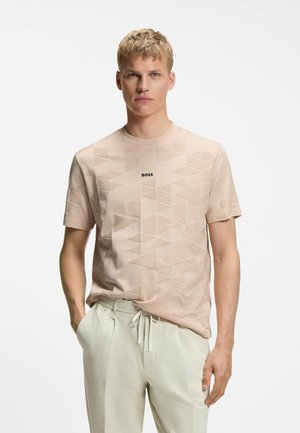 Blondyn ubrany w beżowy t-shirt BOSS z geometrycznym wzorem oraz jasne spodnie ze ściągaczem, stojący z jedną ręką w kieszeni.