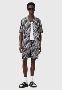 Camicia a maniche corte con bottoni e pantaloni coordinati in un motivo di foglie di palma in bianco e nero. Indossata con una canotta bianca, occhiali da sole neri e sandali.