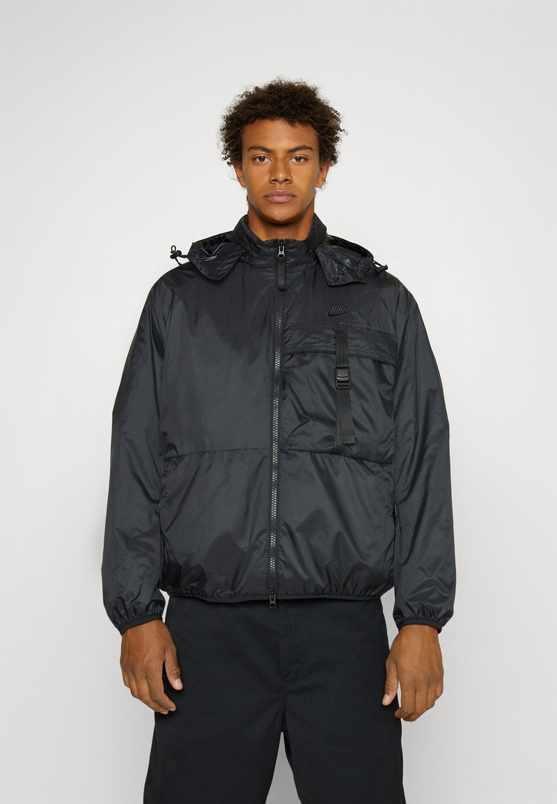 Giacca nera in nylon con zip, cappuccio regolabile, polsini elasticizzati e tasca frontale. Presenta un logo sul petto e un design elegante.