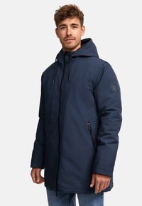 Mørkeblå parka-jakke med hætte, der har lynlås foran, sidelommer og et logo-patch på ærmet. Glat, vandafvisende materiale.
