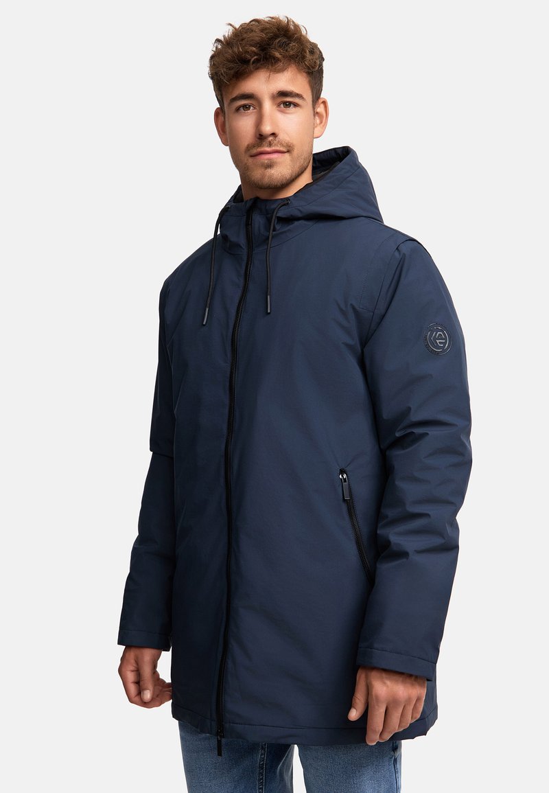 Mørkeblå parka-jakke med hætte, der har lynlås foran, sidelommer og et logo-patch på ærmet. Glat, vandafvisende materiale.