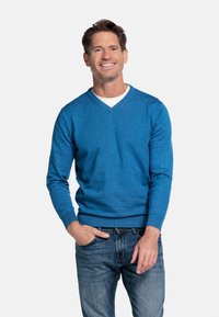 V-hals trui in felblauwe brei, met lange mouwen, geribde manchetten en zoom, gecombineerd met blauwe jeans. Model staat met handen in de zakken.