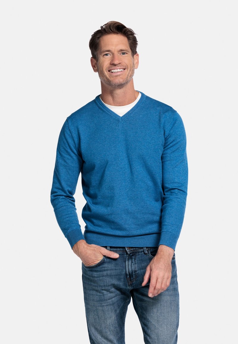 V-hals trui in felblauwe brei, met lange mouwen, geribde manchetten en zoom, gecombineerd met blauwe jeans. Model staat met handen in de zakken.