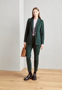 Grön skräddarsydd blazer och matchande byxor i ett glansigt tyg, tillsammans med en ljuslila topp. Svarta klumpiga skor och en brun väska.