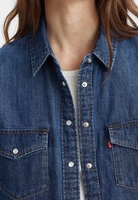 Primo piano di una persona che indossa una camicia blu in denim con bottoni sopra una maglietta bianca a coste, mostrando il colletto, il petto e i bottoni a scatto.