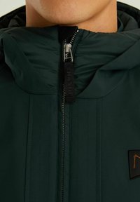 Temno zelena zimska jakna z kapuco, ki ima popoln zadrgo, navpične šive in majhno oznako na prsih.