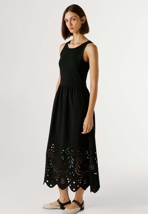 Mujer con vestido midi negro sin mangas con dobladillo ondulado y recortes florales con ojales, combinando sandalias planas beige y negras, de pie.