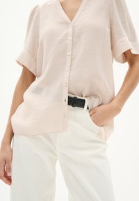 Blusa corta a maniche corte di colore rosa chiaro con scollo a V, chiusura con bottoni e tessuto texture; abbinata a pantaloni bianchi e una cintura nera.