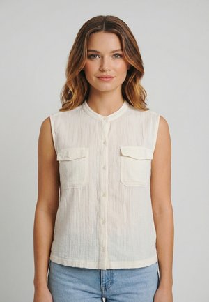 Vrouw met lichtbruin haar draagt een mouwloze witte blouse met knopen en twee borstzakken, en een lichtblauwe spijkerbroek. Ze staat voor een effen achtergrond.