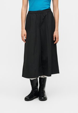 Kvinde iført sorte, vide, cropped bukser, blanke sorte snørestøvler til anklen, hvide sokker og en blå top, stående mod en ensfarvet baggrund.