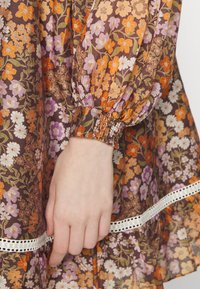 Tecido com padrão floral em tons de castanho, laranja, roxo e branco. Apresenta punhos com elasticidade e um acabamento decorativo em branco.