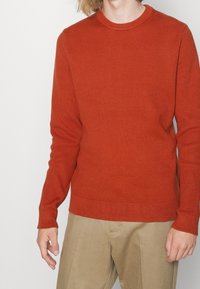 Homme portant un pull ajusté à manches longues orange côtelé et un pantalon beige sur un fond clair uni.