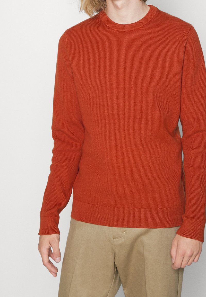 Homme portant un pull ajusté à manches longues orange côtelé et un pantalon beige sur un fond clair uni.