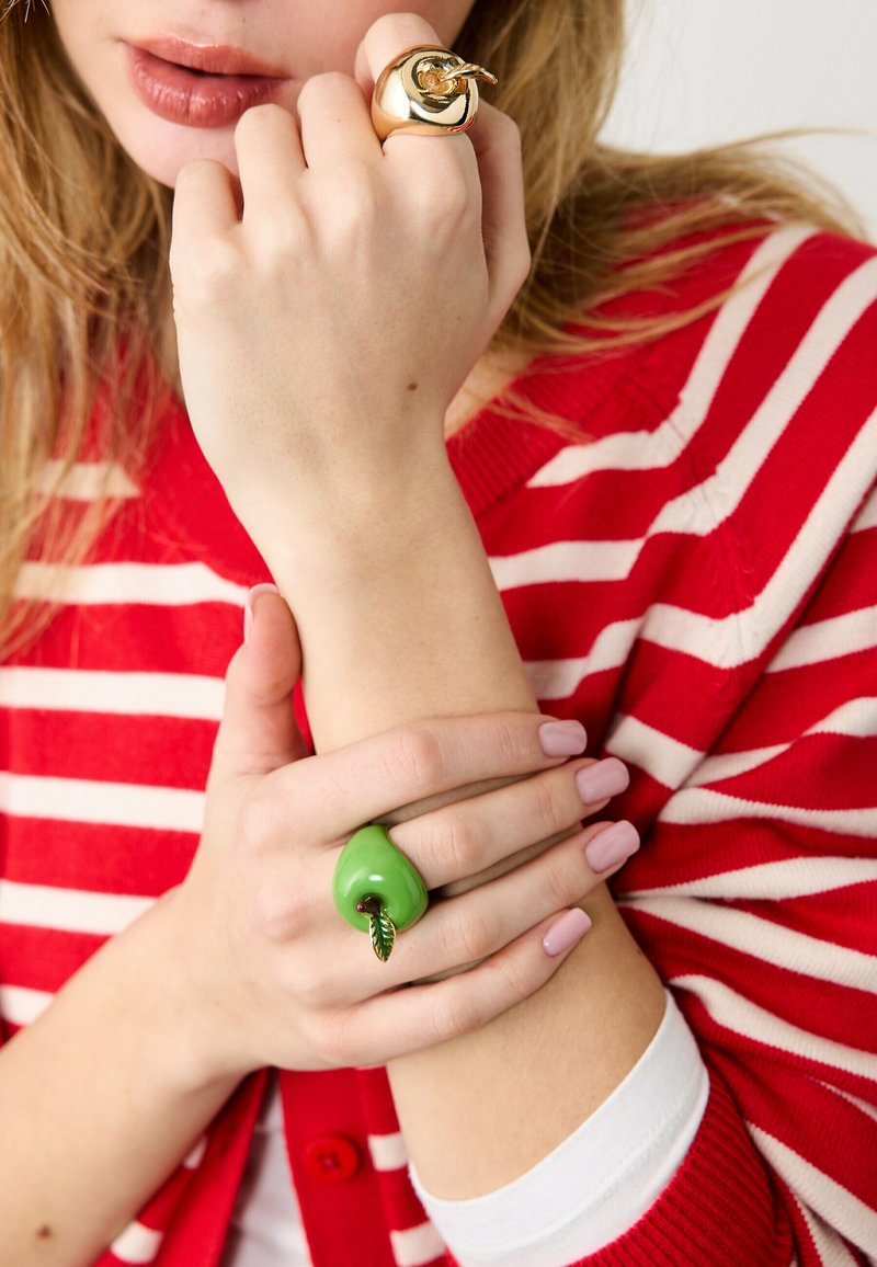Kvinde iført rød- og hvidstribet sweater, der viser begge hænder med store dekorative ringe, den ene guldfarvet og den anden grøn i form af et æble.