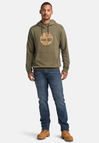 Felpa con cappuccio verde oliva con un grande logo beige a forma di albero, tasca a canguro, cappuccio regolabile con coulisse, abbinata a jeans blu scuro e stivali beige.