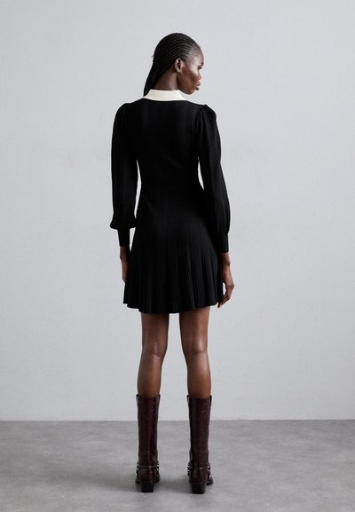 Vestido negro con textura acanalada y falda plisada, mangas largas abullonadas y un collar crema. Combinado con botas burdeos hasta la rodilla con acentos de hardware.