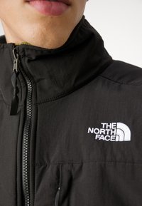 Zwarte jas met ritssluiting en hoge kraag, met een wit logo op de borst, gestructureerde stof en zichtbare rits aan de voorkant.