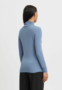 Majica s dolgimi rokavi in turtleneck ovratnikom iz mehkega svetlo modrega blaga, z oblikovanim krojem in subtilnim nabranim detajlom na vratu, v kombinaciji z črnimi hlačami.
