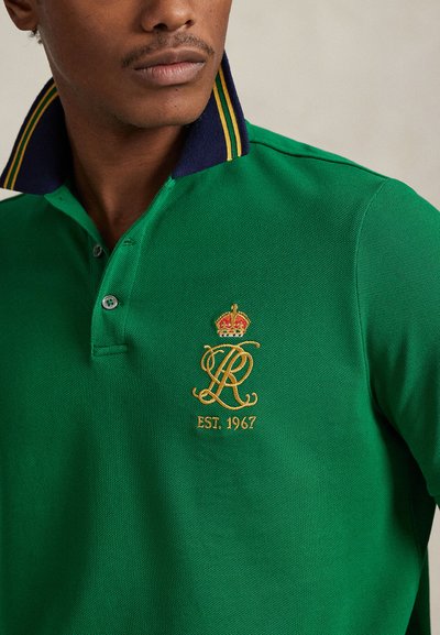 Πράσινο πόλο πουκάμισο κατασκευασμένο από υφασμάτινη υφή, με γιακά σε ναυτικό μπλε χρώμα με κίτρινες ρίγε ς, και κεντητό λογότυπο κορώνας με την ένδειξη "EST. 1967".