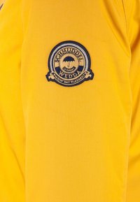 Gul tyg med en släta yta som har en rund, broderad logotyp i blått och svart med ett paraplymotiv.