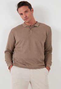Polo shirt beige a maniche lunghe realizzato in tessuto a coste con colletto, due bottoni frontali e polsini aderenti, indossato con pantaloni chiari.