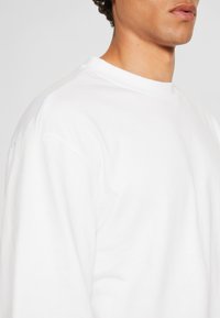 Sweatshirt alb din bumbac, cu guler rotund și mâneci lungi, având o textură netedă și o croială relaxată. Fără logo-uri sau modele vizibile.