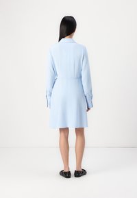 Abito camicia blu chiaro con maniche lunghe, colletto e vita stretta. Realizzato in un tessuto testurizzato, arriva sopra il ginocchio, abbinato a scarpe nere.
