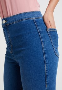 Person wearing blue denim jeans and a pink top with one hand placed inside a back pocket against a plain background.