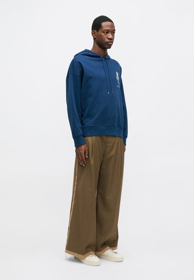 Sudadera con capucha de color azul marino y bordado en la parte frontal, combinada con pantalones de pierna ancha marrones con franjas laterales y zapatillas blancas.