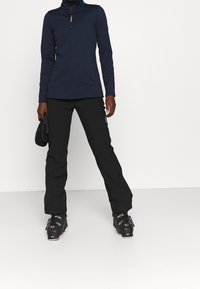 Donna che indossa una giacca con zip blu navy, pantaloni da neve neri, scarponi da sci, con guanti neri in mano su uno sfondo bianco semplice.