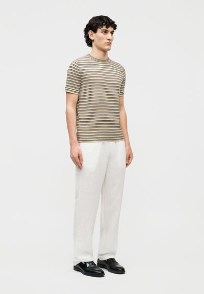 Άνδρας που στέκεται, φοράει μπεζ και μαύρο ριγωτό κοντομάνικο πουκάμισο, λευκό παντελόνι και μαύρα επίσημα παπούτσια, μπροστά από ένα απλό ανοιχτόχρωμο φόντο.