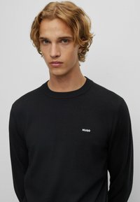 Zwarte crewnecktrui van een zachte gebreide stof, met een klein wit "HUGO" logo op de linkerborst. Eenvoudig, gestroomlijnd ontwerp.