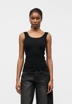 Νεαρή γυναίκα με πλεξούδες, ντυμένη με μαύρο ριγέ τοπ χωρίς μανίκια και σκούρο γκρι τζιν, που στέκεται μπροστά σε απλό λευκό φόντο.